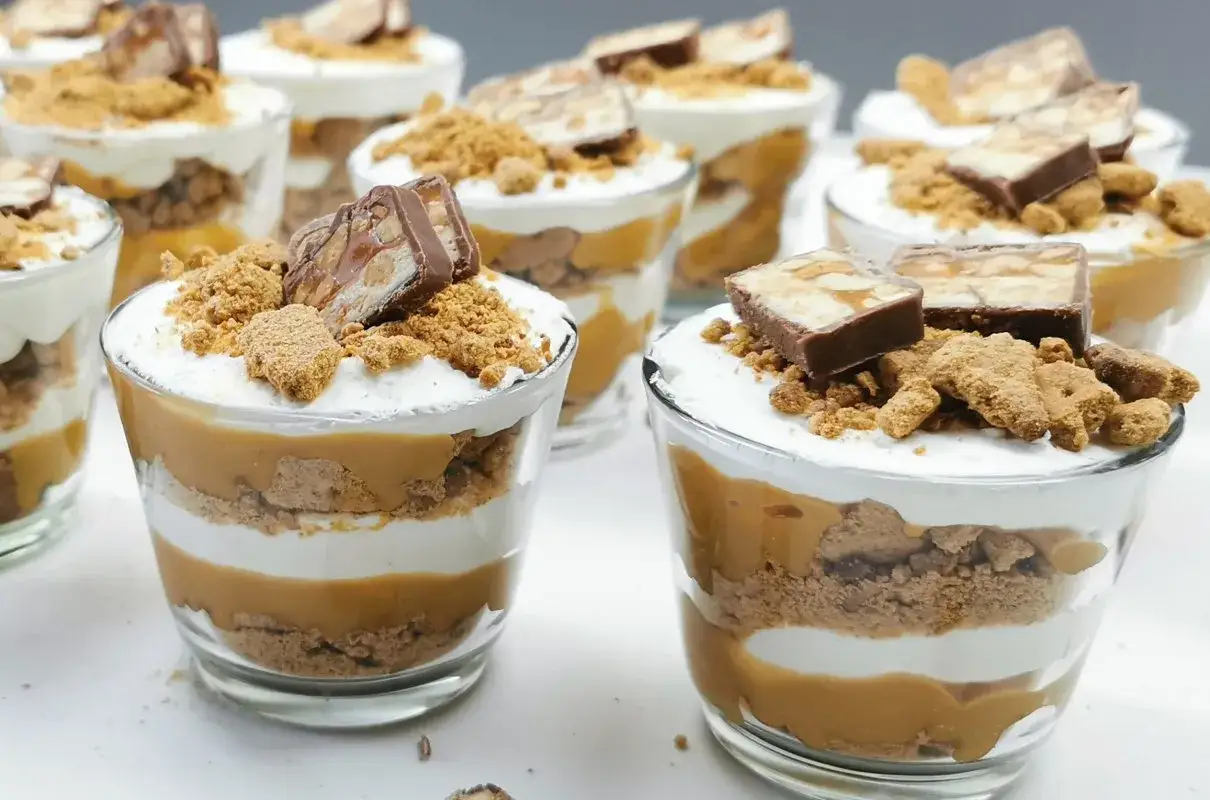 Deserki w pucharkach z warstwami ciastek, kremu i słodkiego dulce de leche, zwieńczone kawałkami batonika.
