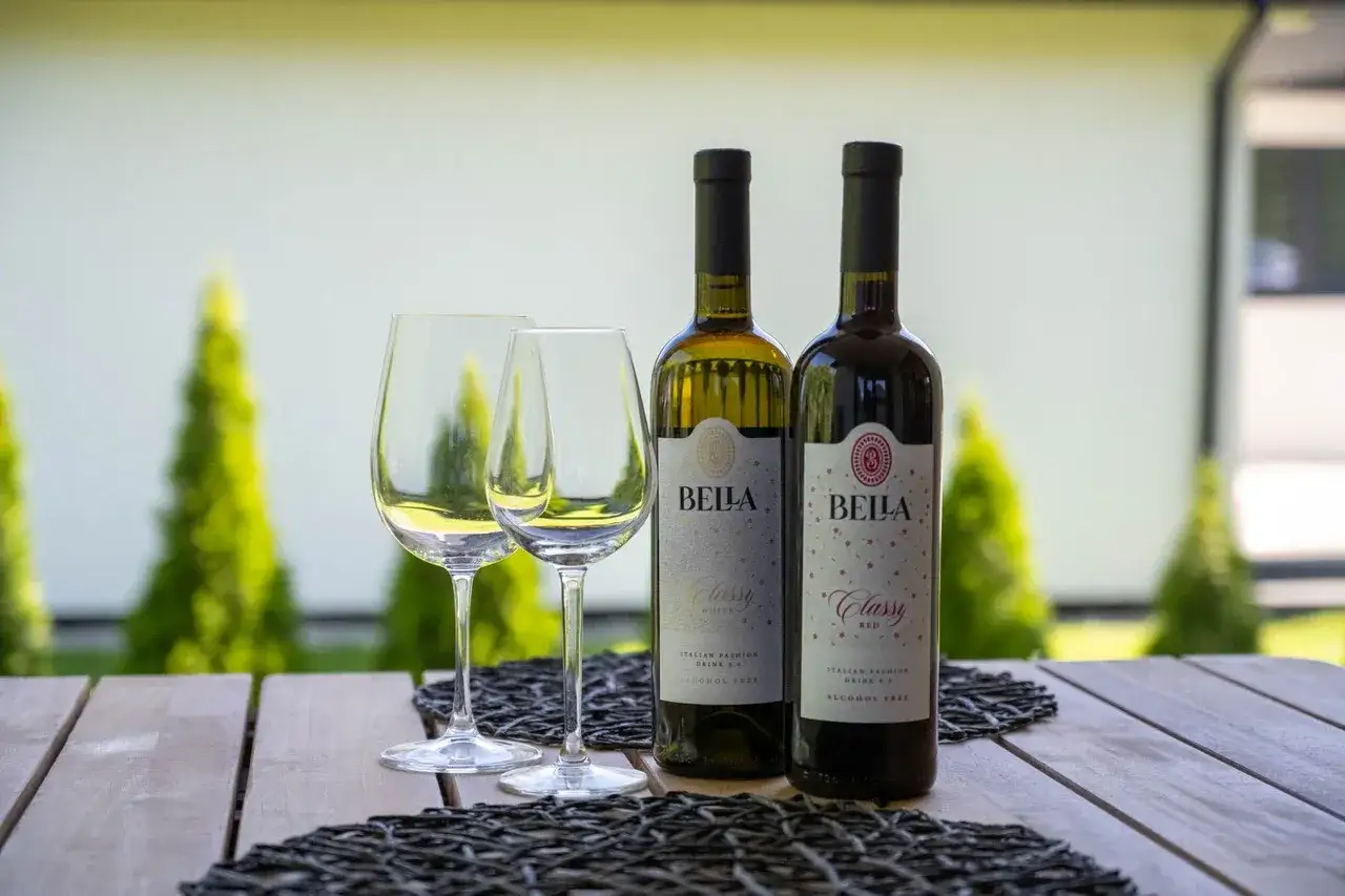 Dwa kieliszki i butelki wina bezalkoholowego Bella Classy White i Red na drewnianym stole.
