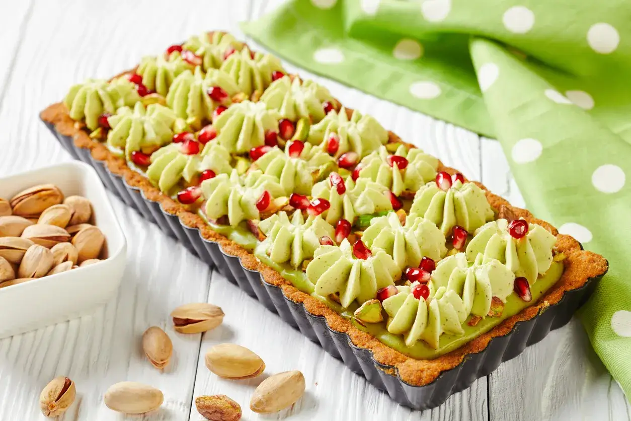 Nowoczesny mazurek wielkanocny z pistacjami i granatem, udekorowany kremem w kształcie rozetek.