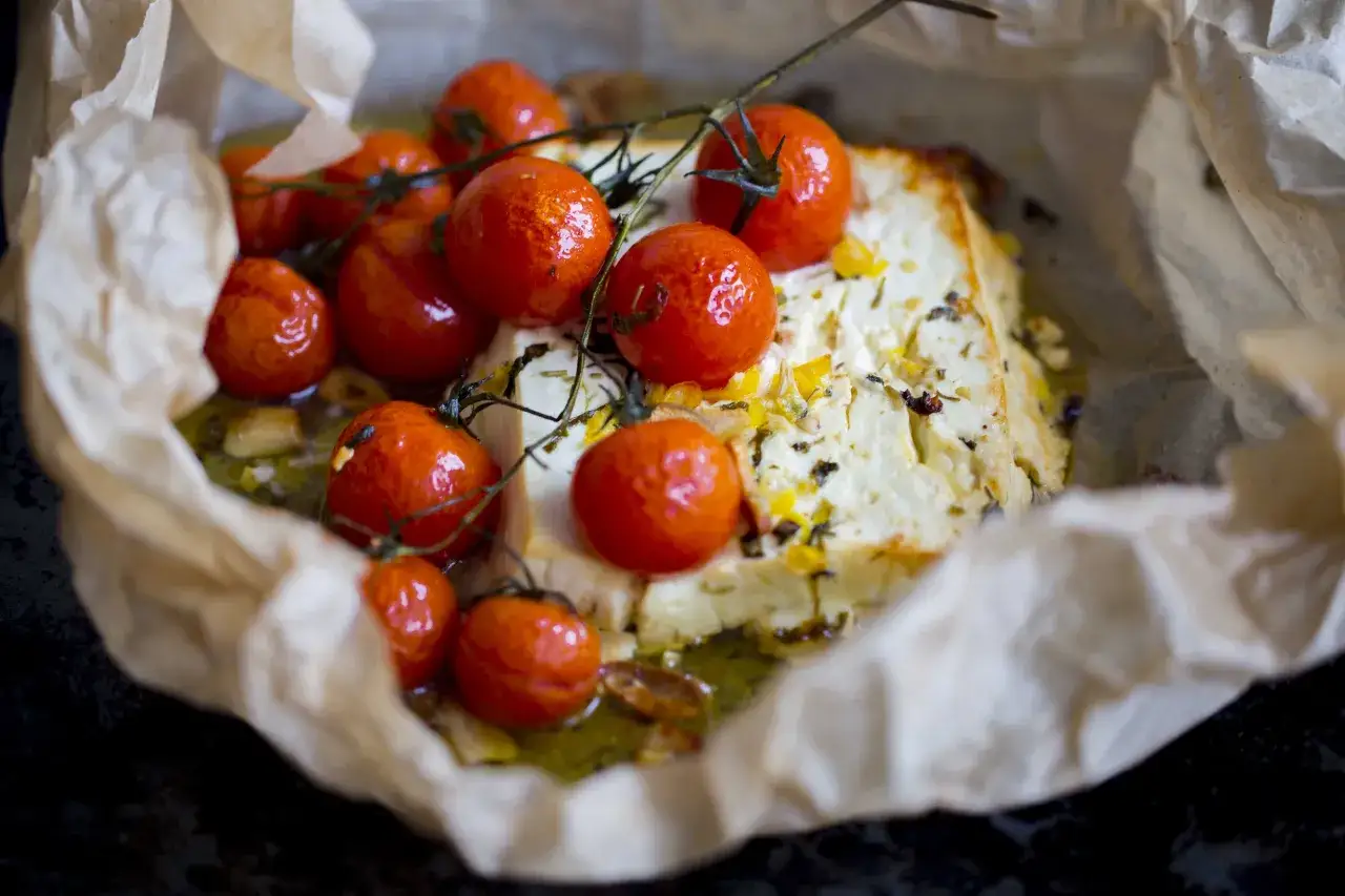 Pieczona feta z pomidorkami koktajlowymi, czosnkiem i ziołami, podana na papierze do pieczenia.