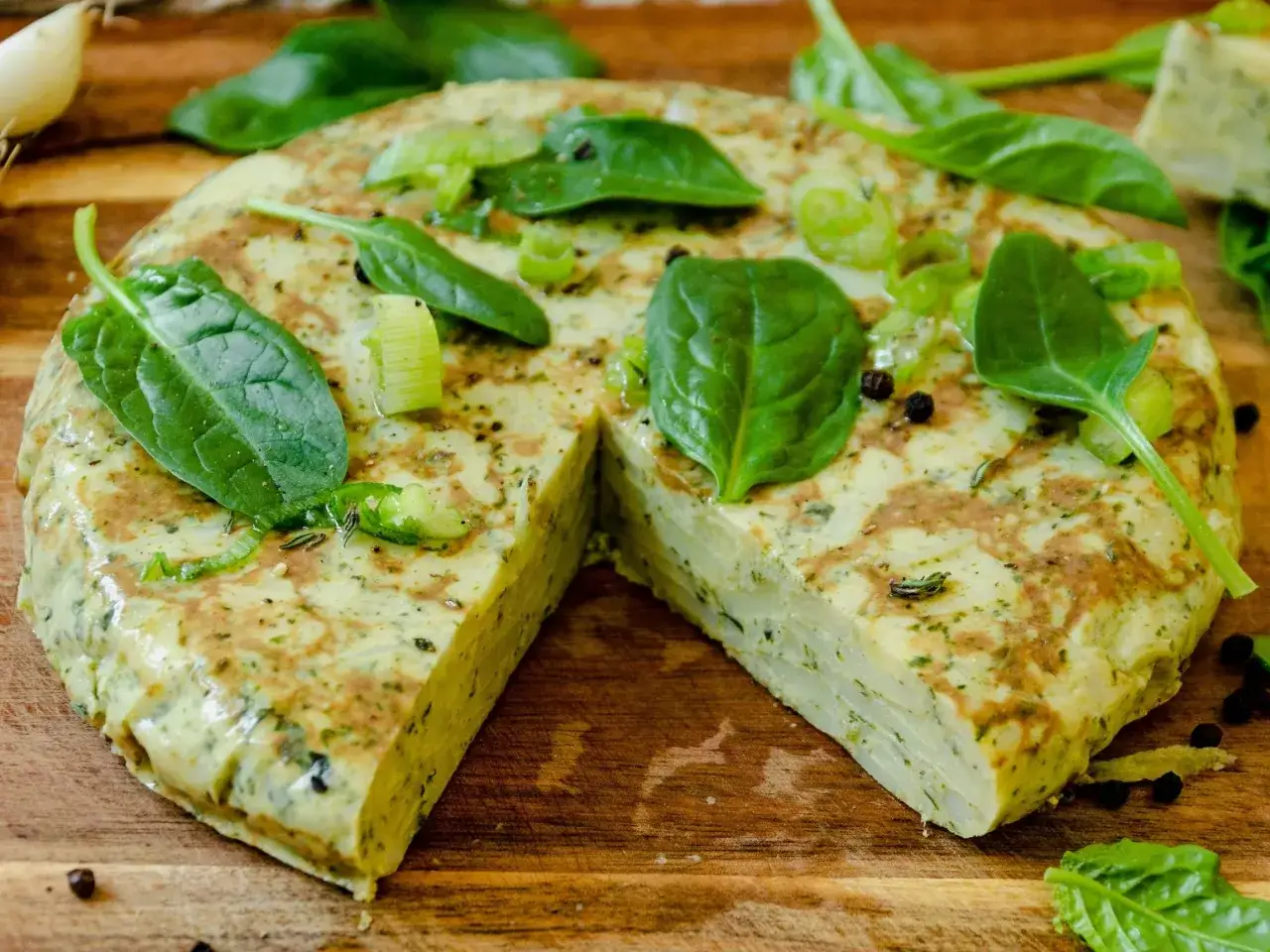 Pyszna tortilla de patatas, grubo pokrojona, ozdobiona świeżym szpinakiem i szczypiorkiem, leży na drewnianej desce.
