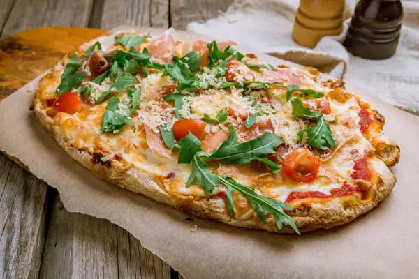 Pyszna pinsa z szynką parmeńską, rukolą i pomidorkami koktajlowymi, posypana parmezanem.