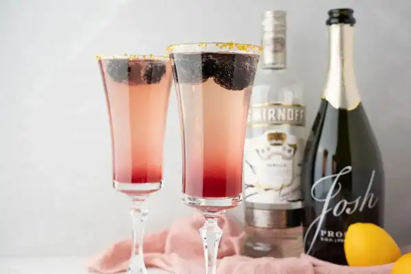 Dwa eleganckie drinki z prosecco, ozdobione jeżynami i złotym cukrem na krawędzi kieliszków. Obok butelka prosecco i wódki.