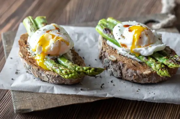 Dwa tosty z pełnoziarnistego pieczywa z jajkiem w koszulce i zielonymi szparagami na śniadanie.