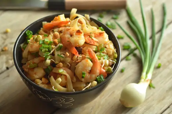 Aksamitne pad thai z krewetkami, posypane szczypiorkiem i orzeszkami, czeka w czarnej misce na drewnianym blacie. Obok leży cebulka.