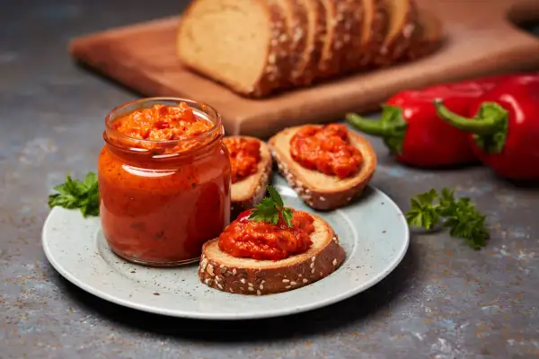 Słoik ajvaru i pieczywo z pastą. Ajvar do czego? Idealny na kanapki, jako dodatek do mięs czy warzyw.