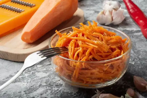 Świeża surówka z marchewki, przygotowana na tarce, czeka w szklanej misce na widelcu. Obok leżą czosnek i papryczka chili.