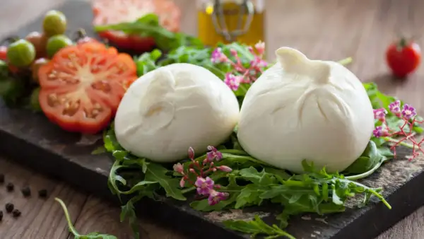 Dwie kulki burraty na rukoli, obok pomidorów i oliwy. To jest burrata – świeży ser z mozzarelli z kremowym wnętrzem.