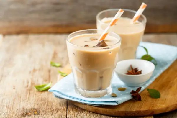 Dwa orzeźwiające **chai latte** z lodem i słomkami, na drewnianym blacie, obok miseczki z anyżem i kardamonem.